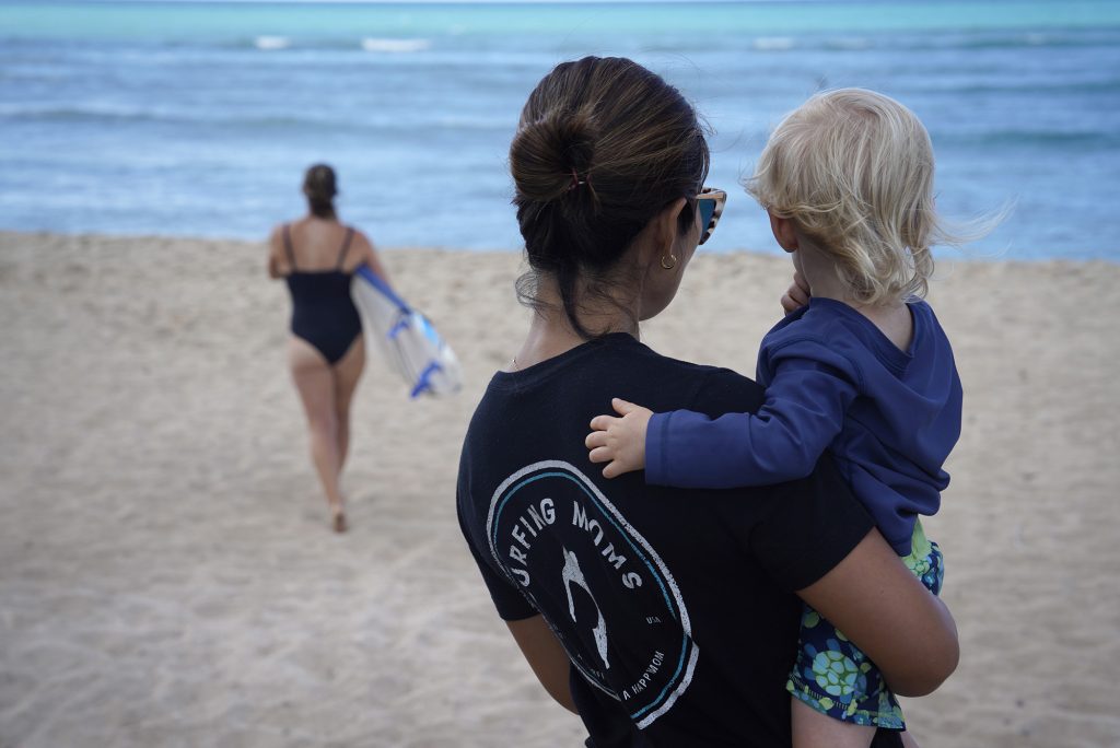 Kelli Ritchie, from left, heads out to get in the water while Sachiko Stokes holds Daniel Ritchie during a Surfing Mom’s weekly outing Tuesday, Aug. 15, 2023, at White Plains Beach in Ewa Beach. The moms take turn surfing while other moms watch the keiki. (Kevin Fujii/Civil Beat/2023)